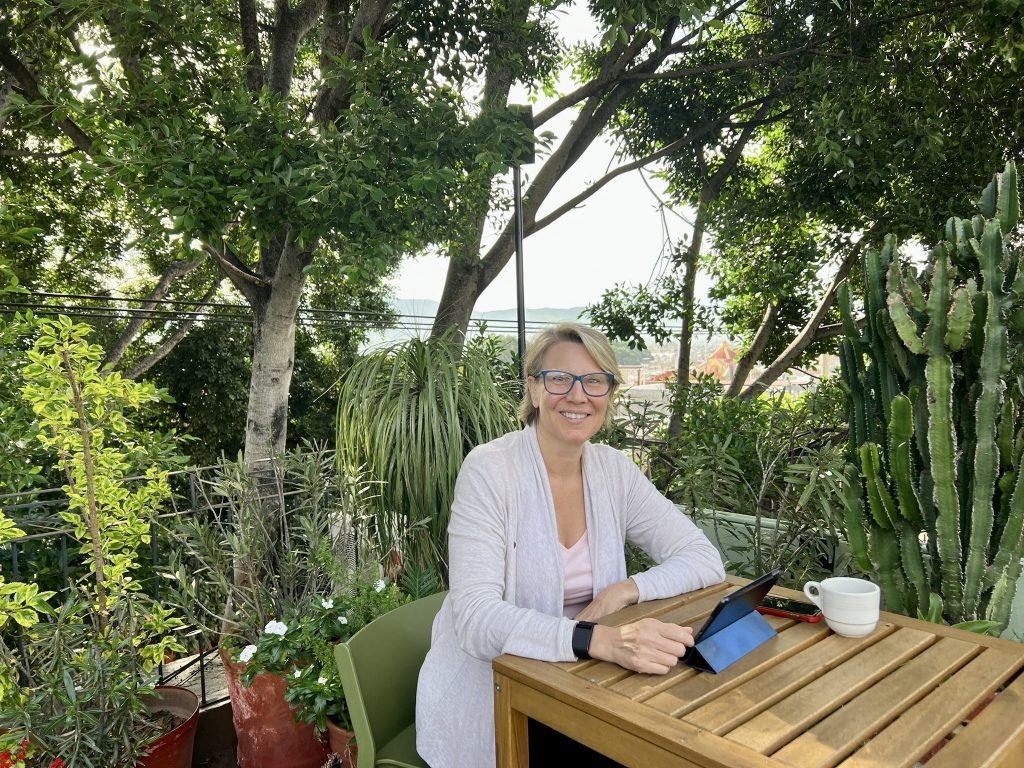The blog writer sitting at a tall table on a rooftop garden, surrounded by green plants. The town of Oaxaca, Mexico, is in the far background.