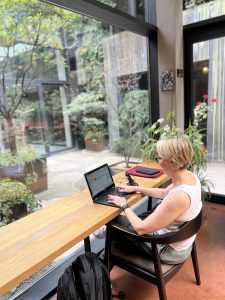 Woman seen from behind working on a laptop on a bench desk in front of a window to a green garden